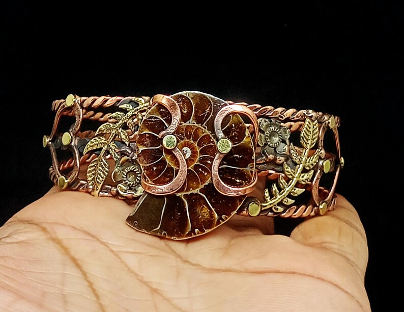 K&ouml;nnte beinhalten: Ein Manschettenarmband aus Kupfer und Messing mit einem gro&szlig;en, braunen Fossilien-Mittelst&uuml;ck. Das Armband hat ein Blumen- und Bl&auml;tterdesign mit einem geflochtenen Seilrand. Kleine gr&uuml;ne Akzente sind sichtbar.
