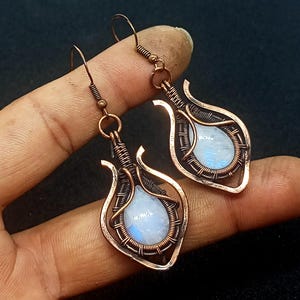 Può includere: Un paio di orecchini avvolti in filo di rame con pietre di luna iridescenti. Gli orecchini hanno la forma di una lacrima con un delicato design in filo.