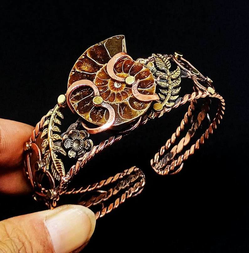 K&ouml;nnte beinhalten: Ein kupferfarbenes Armband mit einem Ammonitenfossil als Mittelst&uuml;ck. Das Armband hat aufwendige Blumen- und Blattdetails mit einem gedrehten Seil-Design am Band. Das Fossil ist eine Mischung aus Braun- und Goldt&ouml;nen mit kleinen Goldakzenten.