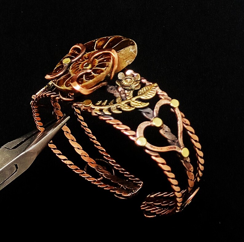 K&ouml;nnte beinhalten: Handgefertigtes Kupferarmband mit filigranen Details. Das Armband zeigt ein Blumenmuster mit goldenen Akzenten, gedrehten, seilartigen Elementen und herzf&ouml;rmigen Ausschnitten. Das Armband ist unten offen und hat einen dunklen Hintergrund.
