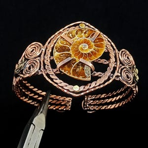 May include: A copper wire-wrapped cuff bracelet featuring a central brown ammonite fossil. The bracelet is constructed with twisted copper wire bands, decorative spiral wire accents on the sides, and small metal studs. A metal tool is visible.