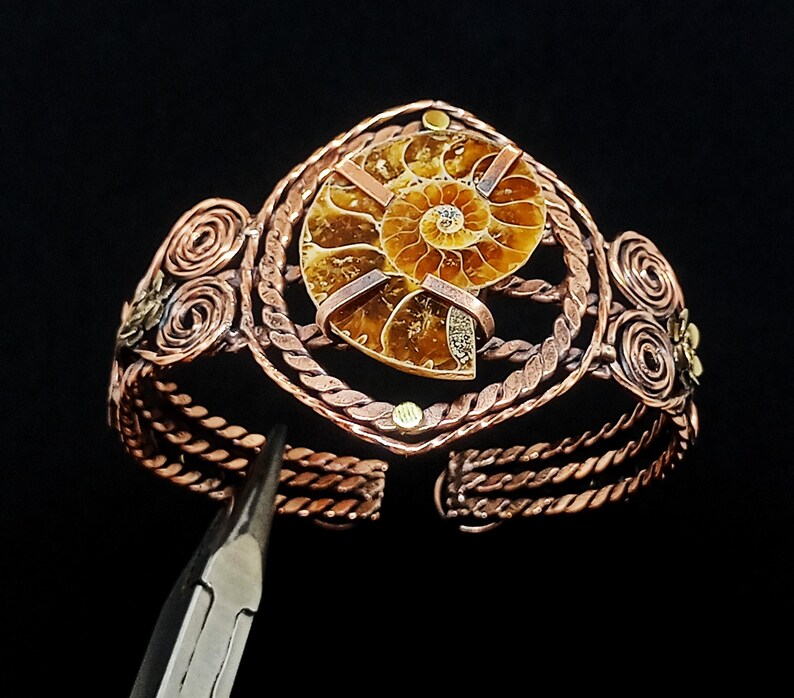 May include: A copper wire-wrapped cuff bracelet featuring a central brown ammonite fossil. The bracelet is constructed with twisted copper wire bands, decorative spiral wire accents on the sides, and small metal studs. A metal tool is visible.
