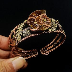 K&ouml;nnte beinhalten: Kupfer-Armreif mit einem Ammoniten-Fossil als Mittelst&uuml;ck. Das Armband hat goldene und silberne Blumen- und Blattdetails sowie ein gedrehtes Seil-Design. Das offene Design erm&ouml;glicht eine verstellbare Passform. Durchmesser ca. 15 cm.