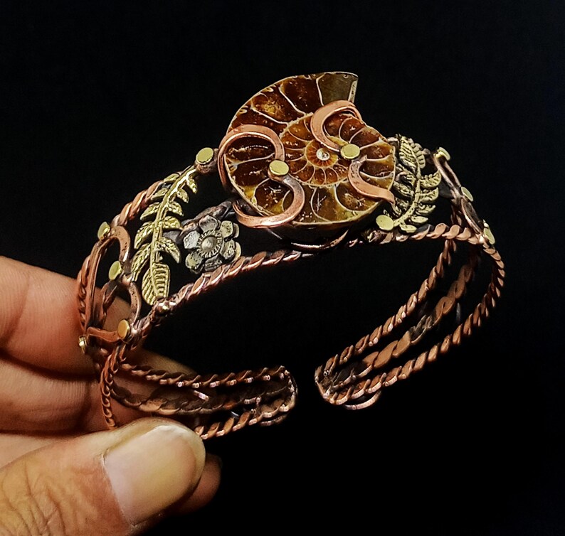 K&ouml;nnte beinhalten: Kupfer-Armreif mit einem Ammoniten-Fossil als Mittelst&uuml;ck. Das Armband hat goldene und silberne Blumen- und Blattdetails sowie ein gedrehtes Seil-Design. Das offene Design erm&ouml;glicht eine verstellbare Passform. Durchmesser ca. 15 cm.
