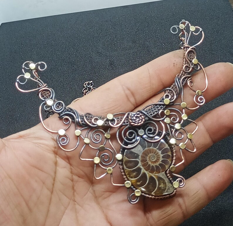 K&ouml;nnte beinhalten: Eine Halskette aus Silber- und Kupferdraht mit einem gro&szlig;en Ammoniten-Fossil als Anh&auml;nger. Die Halskette ist mit kleinen goldenen Perlen verziert und hat ein filigranes, kompliziertes Design.