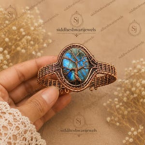 May include: A copper wire-wrapped cuff bracelet featuring an oval labradorite gemstone with a blue iridescent flash. The stone is encased in a detailed wire tree design, with the surrounding band constructed from a woven copper wire lattice pattern.