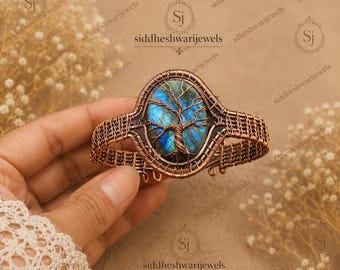 May include: A copper wire-wrapped cuff bracelet featuring an oval labradorite gemstone with a blue iridescent flash. The stone is encased in a detailed wire tree design, with the surrounding band constructed from a woven copper wire lattice pattern.