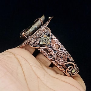 May include: A copper cuff bracelet featuring intricate wire-wrapped scrollwork, twisted metal accents, and a small brass flower detail. A large, textured, dark-coloured stone is securely mounted at the top of the band, creating a bold focal point.