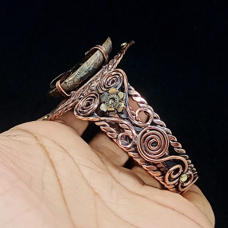 May include: A copper cuff bracelet featuring intricate wire-wrapped scrollwork, twisted metal accents, and a small brass flower detail. A large, textured, dark-coloured stone is securely mounted at the top of the band, creating a bold focal point.