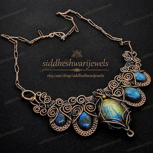 May include: A copper wire-wrapped necklace featuring five iridescent blue and gold labradorite gemstones. The metalwork consists of intricate swirls and loops connected to a chain with a hook clasp. The text siddheshwarijewels is visible.