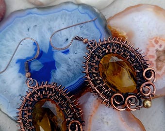 May include: A pair of copper wire wrapped earrings with amber glass stones. The earrings are oval shaped and have a delicate wire design.
