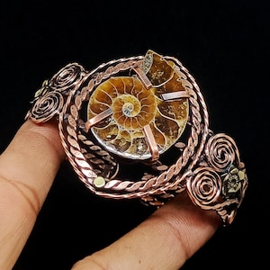 May include: A copper wire cuff bracelet featuring a central spiral ammonite fossil held by metal prongs. The band is constructed from twisted copper wire and decorated with multiple copper wire swirls and small metallic floral accents on the sides.