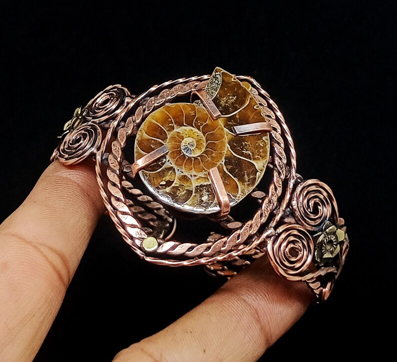 May include: A copper wire cuff bracelet featuring a central spiral ammonite fossil held by metal prongs. The band is constructed from twisted copper wire and decorated with multiple copper wire swirls and small metallic floral accents on the sides.