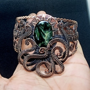 Peut inclure: Un bracelet artisanal en fil de cuivre avec un motif de pieuvre. Le bracelet comporte une pierre verte ovale au centre. Les tentacules de la pieuvre sont finement tissés, et le bracelet a un aspect artistique détaillé.