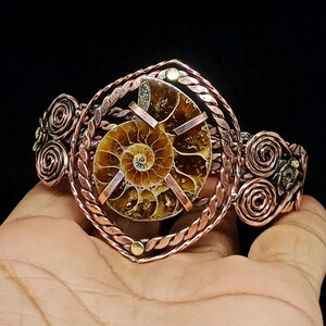 May include: A copper cuff bracelet featuring a central brown and tan spiral ammonite fossil. The fossil is held by three copper prongs within a twisted copper wire frame. The sides of the bracelet are decorated with copper wire swirls.