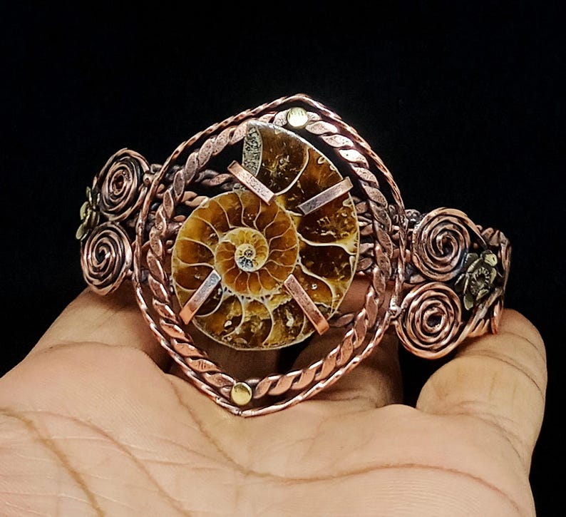 May include: A copper cuff bracelet featuring a central brown and tan spiral ammonite fossil. The fossil is held by three copper prongs within a twisted copper wire frame. The sides of the bracelet are decorated with copper wire swirls.