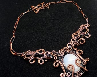 May include: A handcrafted copper wire necklace with an oval, white gemstone pendant. The necklace features intricate wirework with swirling patterns and a chain closure. The jewelry is displayed on a black surface.