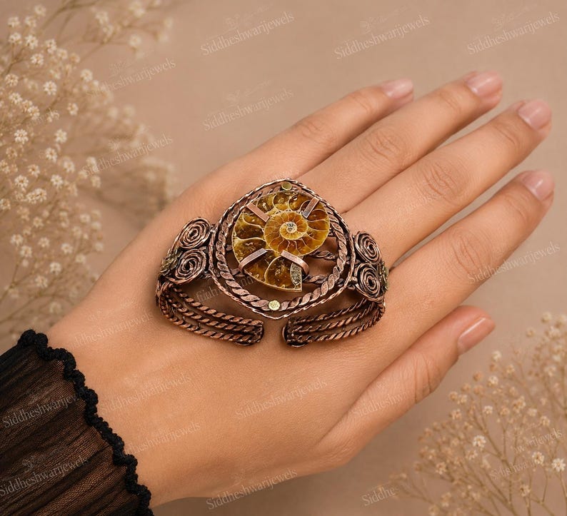 May include: A copper-toned cuff bracelet featuring a central spiral ammonite fossil. The metal band is crafted with twisted wire detailing and decorative coiled accents on both sides. The fossil displays natural brown and tan tones.