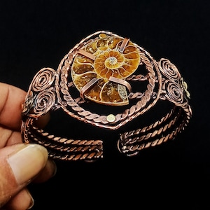May include: A copper cuff bracelet featuring a central brown and tan spiral ammonite fossil. The metal band is constructed from twisted copper wire with decorative coiled wire accents on the sides, securing the fossil with flat copper strips.