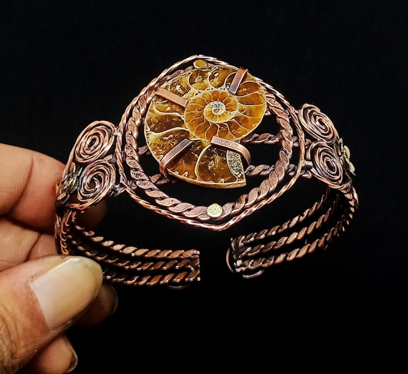 May include: A copper cuff bracelet featuring a central brown and tan spiral ammonite fossil. The metal band is constructed from twisted copper wire with decorative coiled wire accents on the sides, securing the fossil with flat copper strips.