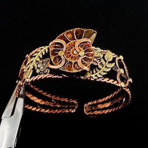 K&ouml;nnte beinhalten: Ein handgefertigtes Kupfer-Armreif-Armband mit einem Ammoniten-Fossil-Mittelst&uuml;ck. Das Armband hat aufwendige Blumen- und Blattdetails in Gold- und Bronzet&ouml;nen. Das offene Design erm&ouml;glicht eine verstellbare Passform. Der Hintergrund ist schwarz.