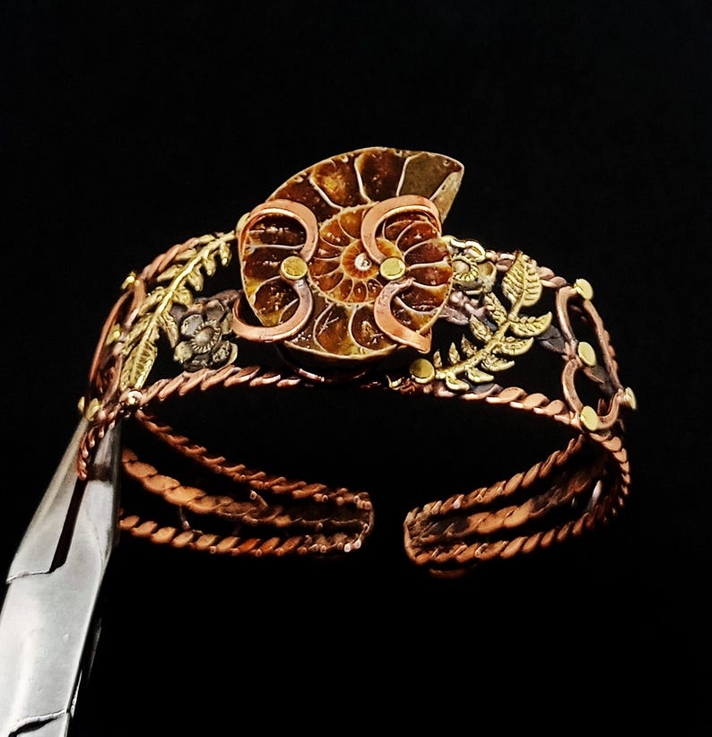 K&ouml;nnte beinhalten: Ein handgefertigtes Kupfer-Armreif-Armband mit einem Ammoniten-Fossil-Mittelst&uuml;ck. Das Armband hat aufwendige Blumen- und Blattdetails in Gold- und Bronzet&ouml;nen. Das offene Design erm&ouml;glicht eine verstellbare Passform. Der Hintergrund ist schwarz.