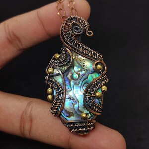 May include: A pendant featuring an iridescent abalone shell center encased in intricate, dark-toned copper wire wrapping. The design includes small, round, metallic gold-colored beads integrated into the wirework and a spiral top detail.