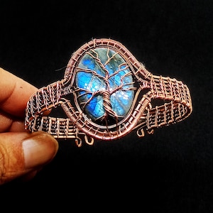 May include: Copper wire cuff bracelet with a central oval blue gemstone and a tree design. The bracelet has an intricate woven wire design. The gemstone has a blue and green iridescent effect. The bracelet is held by a hand against a black background.
