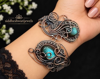 Puede incluir: Un brazalete de alambre de cobre envuelto con dos piedras preciosas de labradorita ovaladas con iridiscencia azul. El trabajo en metal consiste en intrincados remolinos y patrones tejidos, creando un marco detallado alrededor de cada piedra. El texto siddheshwarijewels es visible.