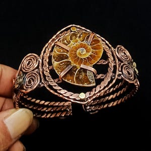May include: A copper cuff bracelet featuring a central brown ammonite fossil encased in a twisted wire frame. The band is constructed from multiple strands of braided copper wire with decorative spiral accents on the sides and small metal studs.