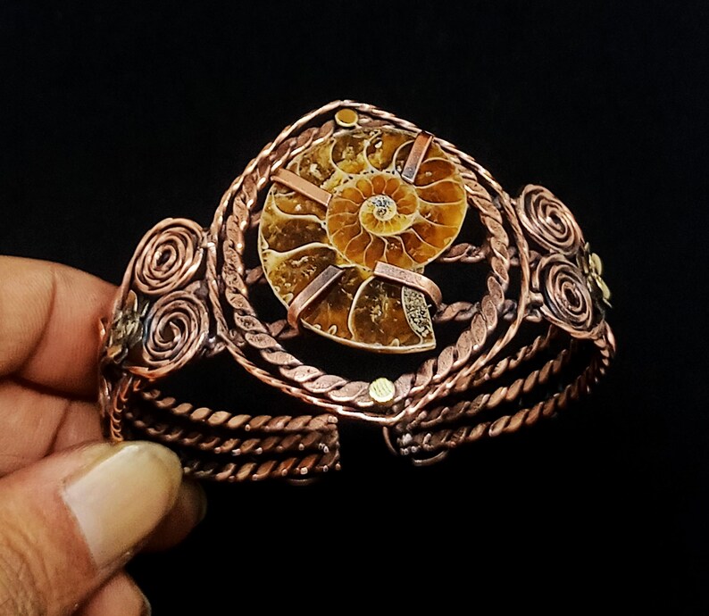 May include: A copper cuff bracelet featuring a central brown ammonite fossil encased in a twisted wire frame. The band is constructed from multiple strands of braided copper wire with decorative spiral accents on the sides and small metal studs.