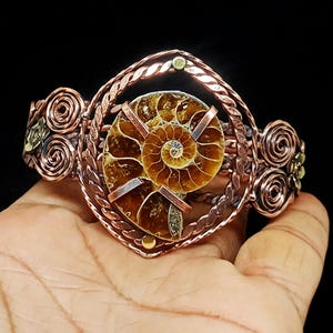 May include: A copper wire-wrapped cuff bracelet featuring a central brown spiral ammonite fossil. The metalwork includes twisted wire borders and decorative circular wire coils on both sides, creating a detailed, structured metallic accessory.