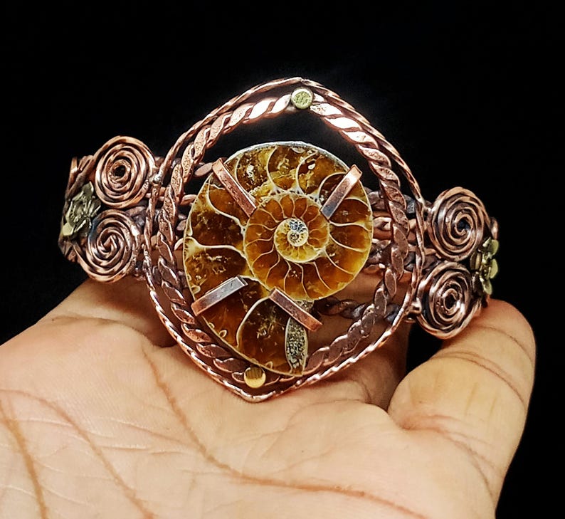 May include: A copper wire-wrapped cuff bracelet featuring a central brown spiral ammonite fossil. The metalwork includes twisted wire borders and decorative circular wire coils on both sides, creating a detailed, structured metallic accessory.