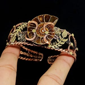 K&ouml;nnte beinhalten: Ein kupferfarbenes Armband mit einem Ammonitenfossil als Mittelst&uuml;ck. Das offene Design umfasst florale und Blattakzente in Gold- und Silbert&ouml;nen. Das Armband hat eine gedrehte Seiltextur.