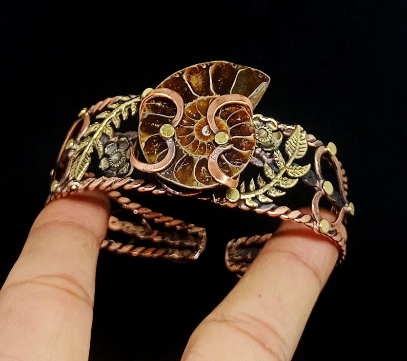 K&ouml;nnte beinhalten: Ein kupferfarbenes Armband mit einem Ammonitenfossil als Mittelst&uuml;ck. Das offene Design umfasst florale und Blattakzente in Gold- und Silbert&ouml;nen. Das Armband hat eine gedrehte Seiltextur.
