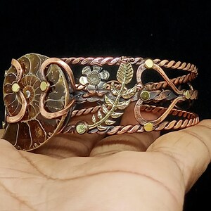 K&ouml;nnte beinhalten: Ein handgefertigtes Kupferarmband mit einem Ammonitenfossil, floralen Akzenten und einem Blattdesign. Das Armband hat ein gedrehtes Seildesign und ist mit goldfarbenen Nieten verziert. Das Armband ist etwa 5 cm breit.
