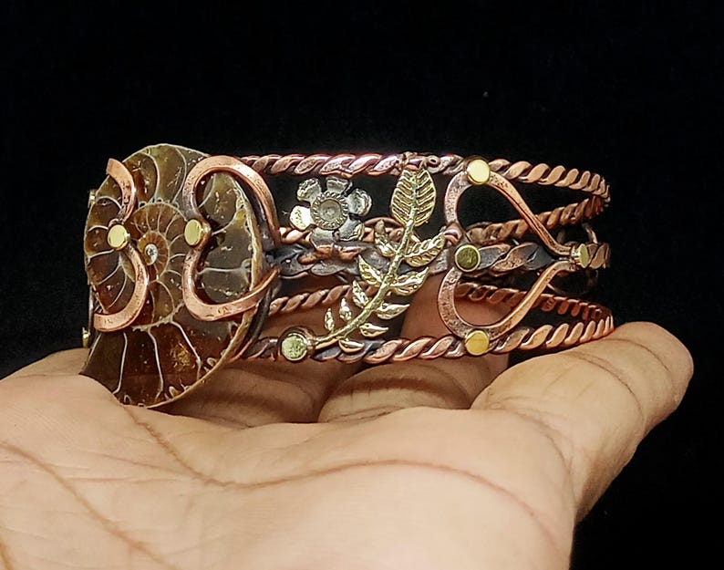 K&ouml;nnte beinhalten: Ein handgefertigtes Kupferarmband mit einem Ammonitenfossil, floralen Akzenten und einem Blattdesign. Das Armband hat ein gedrehtes Seildesign und ist mit goldfarbenen Nieten verziert. Das Armband ist etwa 5 cm breit.