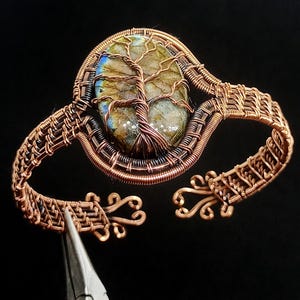 May include: Copper wire cuff bracelet with an oval stone centre, featuring a tree of life design. The stone has green, blue, and gold hues. The bracelet has an open design with detailed wirework and decorative swirls.