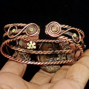 K&ouml;nnte beinhalten: Kupfer-Armreif mit aufwendigen Blumen- und Spiralmustern. Das offene Armband hat einen gedrehten Seilrand und kleine dekorative Akzente. Das Metall hat einen warmen, r&ouml;tlich-braunen Farbton mit goldenen und gr&uuml;nen Akzenten.