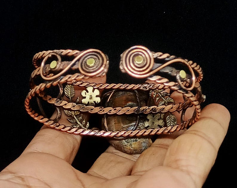 K&ouml;nnte beinhalten: Kupfer-Armreif mit aufwendigen Blumen- und Spiralmustern. Das offene Armband hat einen gedrehten Seilrand und kleine dekorative Akzente. Das Metall hat einen warmen, r&ouml;tlich-braunen Farbton mit goldenen und gr&uuml;nen Akzenten.