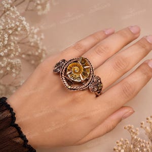 May include: A copper-toned ring featuring a central spiral ammonite fossil. The metal band is crafted with intricate wire-wrapped detailing, including twisted rope patterns and decorative coiled accents on both sides of the fossil setting.