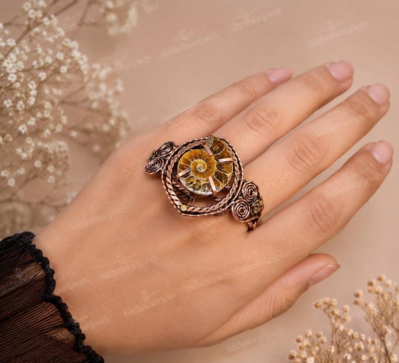 May include: A copper-toned ring featuring a central spiral ammonite fossil. The metal band is crafted with intricate wire-wrapped detailing, including twisted rope patterns and decorative coiled accents on both sides of the fossil setting.