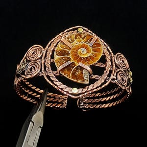 May include: A copper wire-wrapped cuff bracelet featuring a central spiral ammonite fossil. The bracelet is constructed from twisted copper wire strands, decorative wire coils, and small brass accents, with a pair of metal pliers visible.