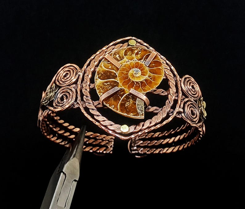 May include: A copper wire-wrapped cuff bracelet featuring a central spiral ammonite fossil. The bracelet is constructed from twisted copper wire strands, decorative wire coils, and small brass accents, with a pair of metal pliers visible.