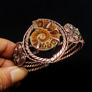 May include: A cuff bracelet crafted from twisted copper wire featuring a central brown ammonite fossil. The design includes decorative wire spirals on the sides and metal accents, with the fossil secured by thin copper bands across its surface.