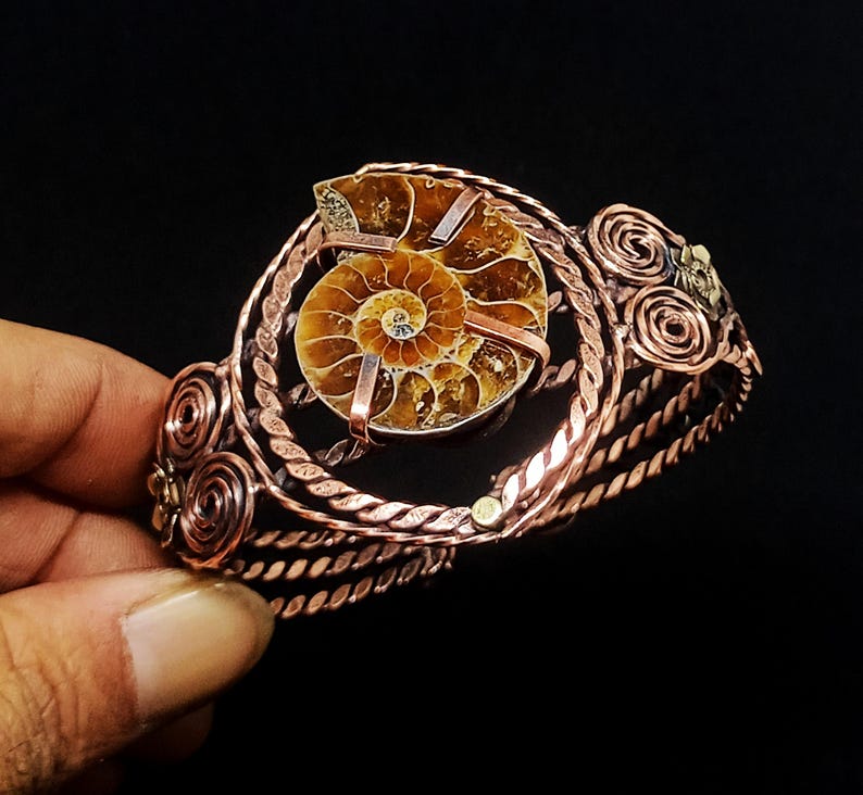 May include: A cuff bracelet crafted from twisted copper wire featuring a central brown ammonite fossil. The design includes decorative wire spirals on the sides and metal accents, with the fossil secured by thin copper bands across its surface.