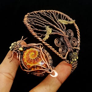 Könnte beinhalten: Ein handgefertigtes Kupferdrahtarmband mit einem Fossilanhänger. Das Armband hat ein Baumdesign mit Messingvögeln und floralen Akzenten. Der Anhänger zeigt ein spiralförmiges Fossil in Orange- und Brauntönen.