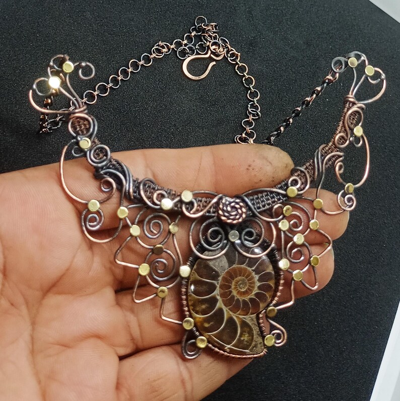 K&ouml;nnte beinhalten: Eine Halskette aus Kupfer- und Silberdraht mit einem gro&szlig;en Ammoniten-Fossil-Anh&auml;nger. Die Halskette hat ein filigranes, kompliziertes Design mit kleinen goldenen Perlen.