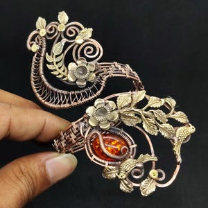 May include: Ornate copper-toned bracelet with floral and leaf accents. Features a central amber-colored stone and intricate wirework. Includes brass-colored flowers and bird details, set against a dark background.