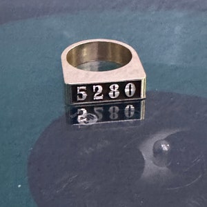 May include: A silver ring with a rectangular band featuring the numbers "5280" in black.